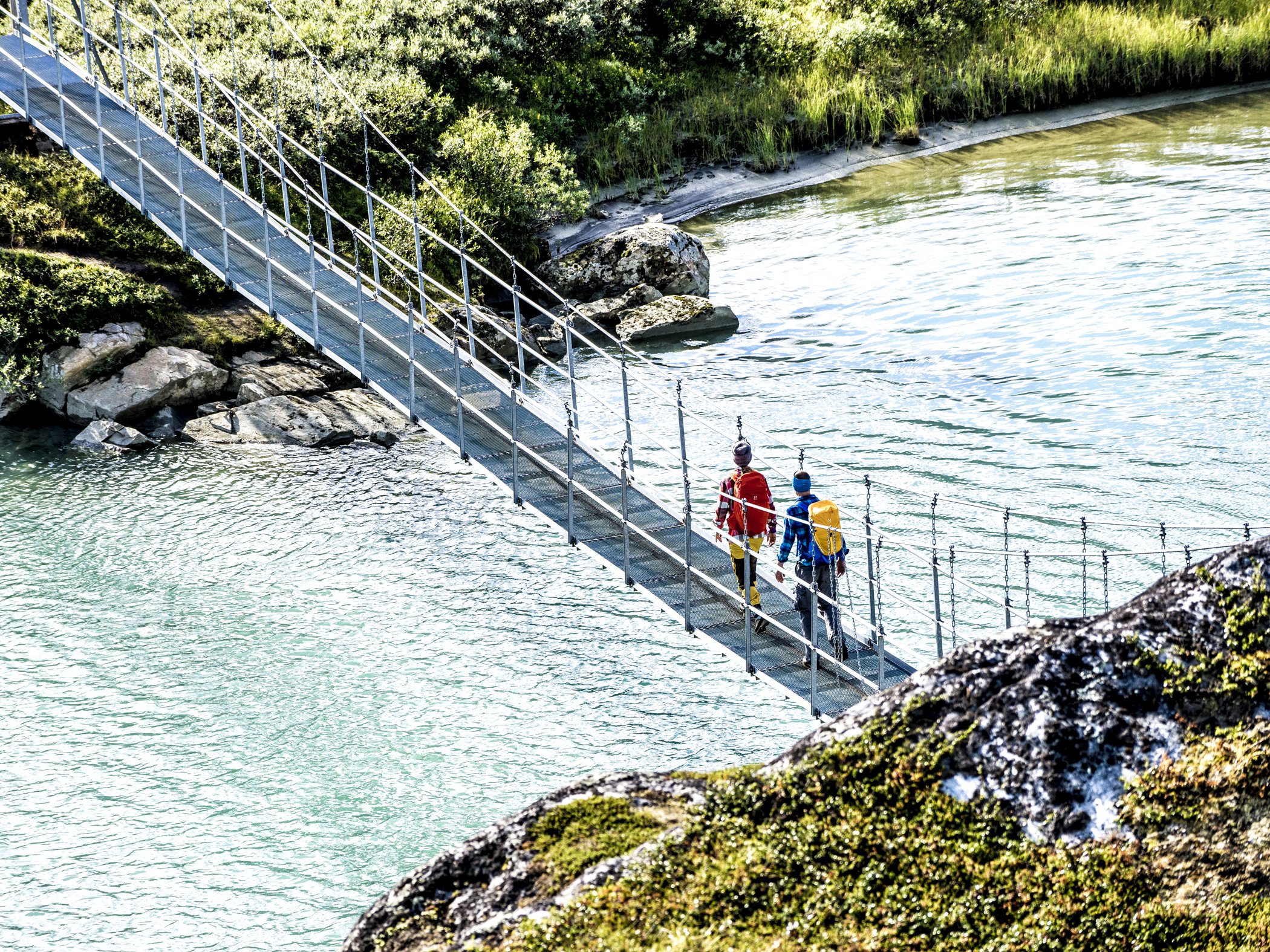Hikers crossing a suspension bridge over turquoise water.