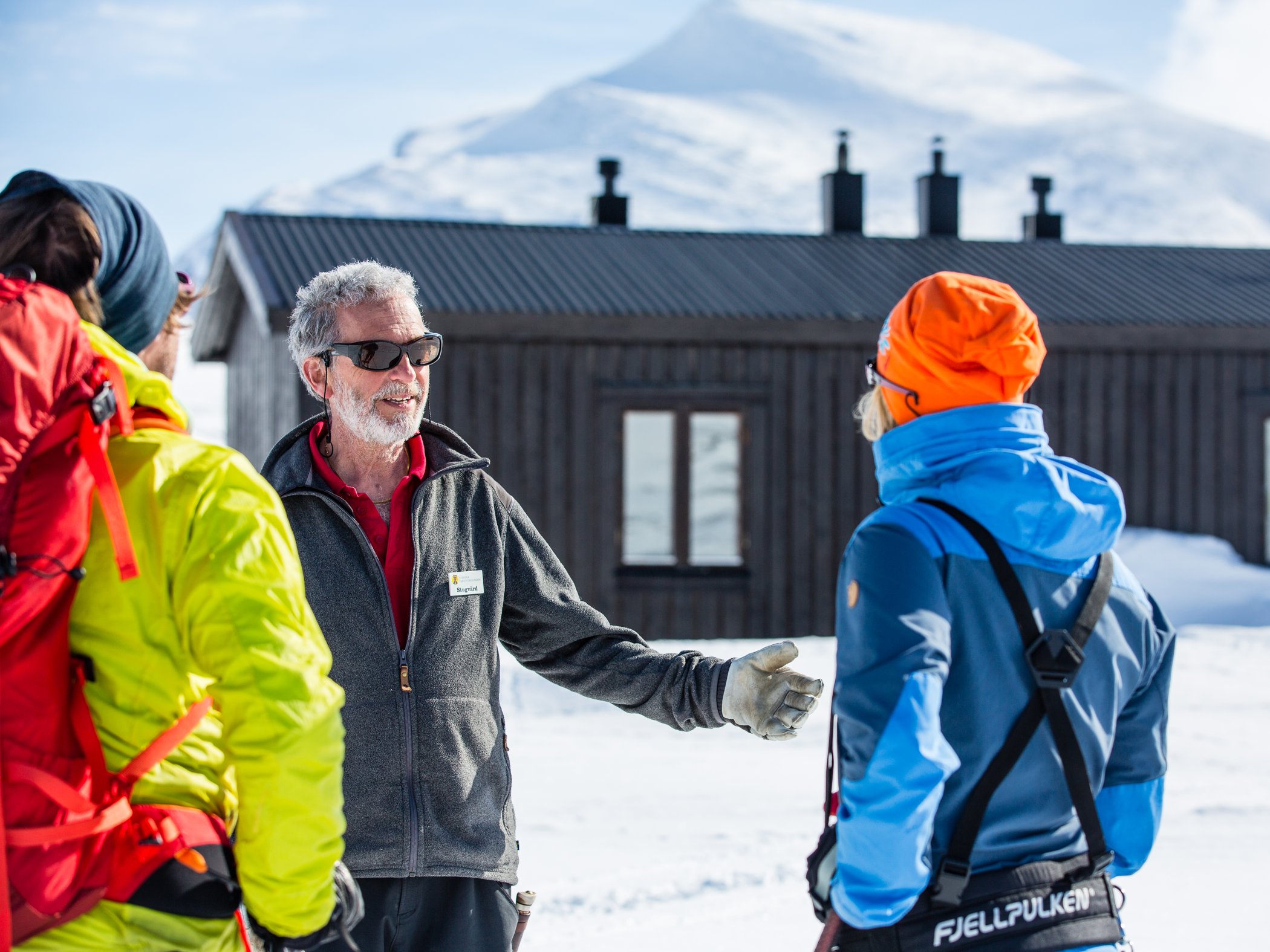 The cabin host welcomes two skiers outside the mountain cabin on a winter day.