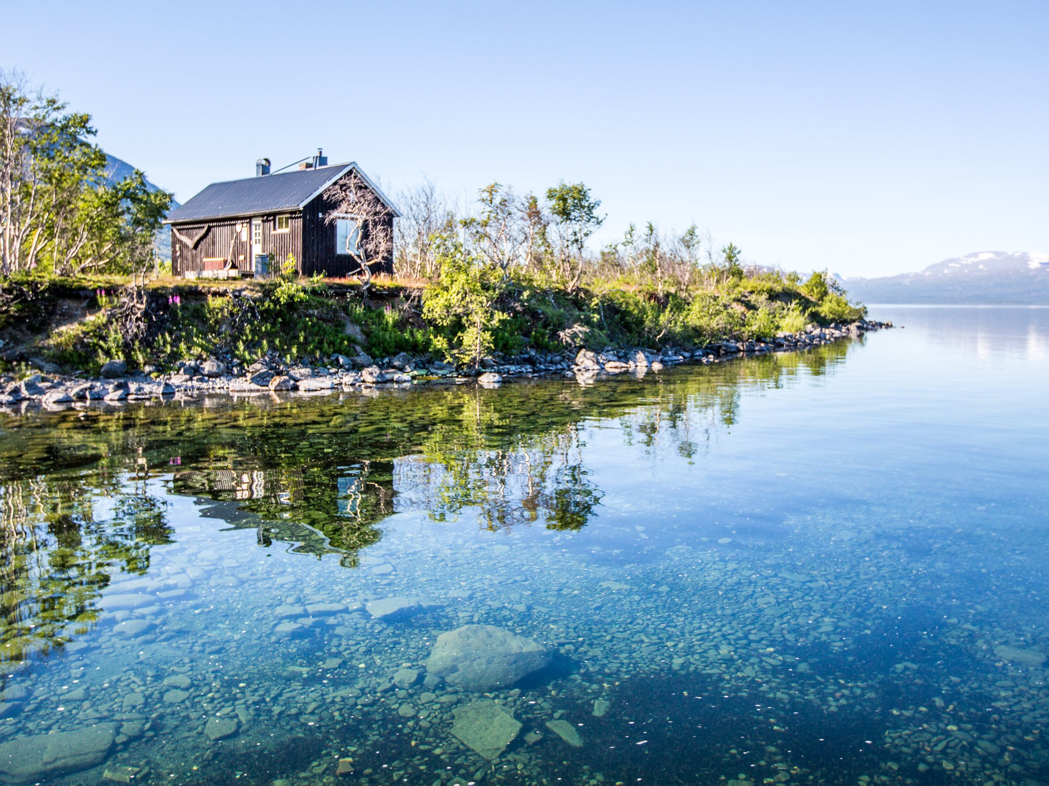 Sauna by the shore of Lake Torneträsk with clear water and mountains in the background.