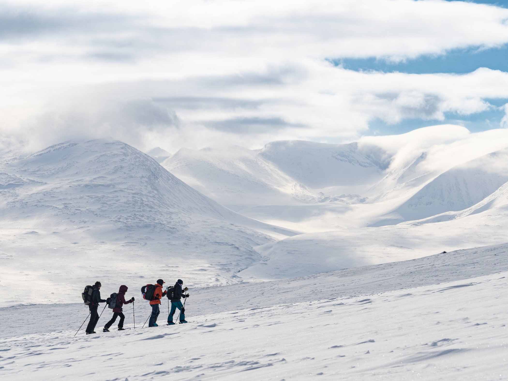 Four skiers in the distance walking in single file through a white snowy landscape.