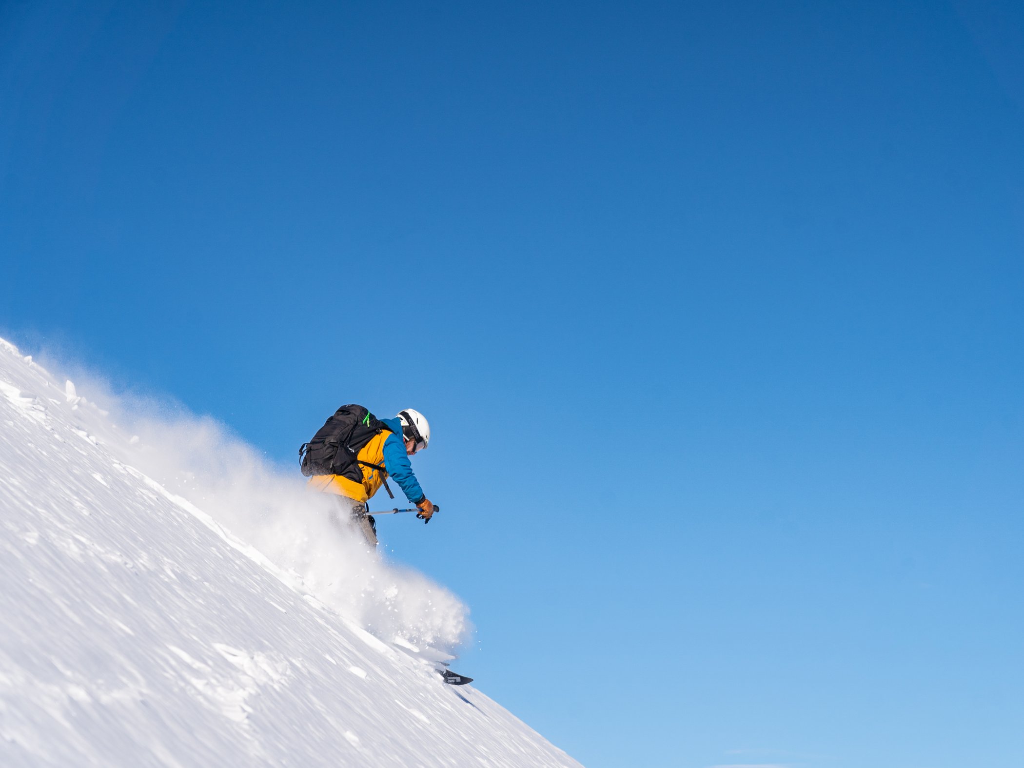 A skier in a yellow and blue jacket skiing down a steep mountain slope under a blue sky.