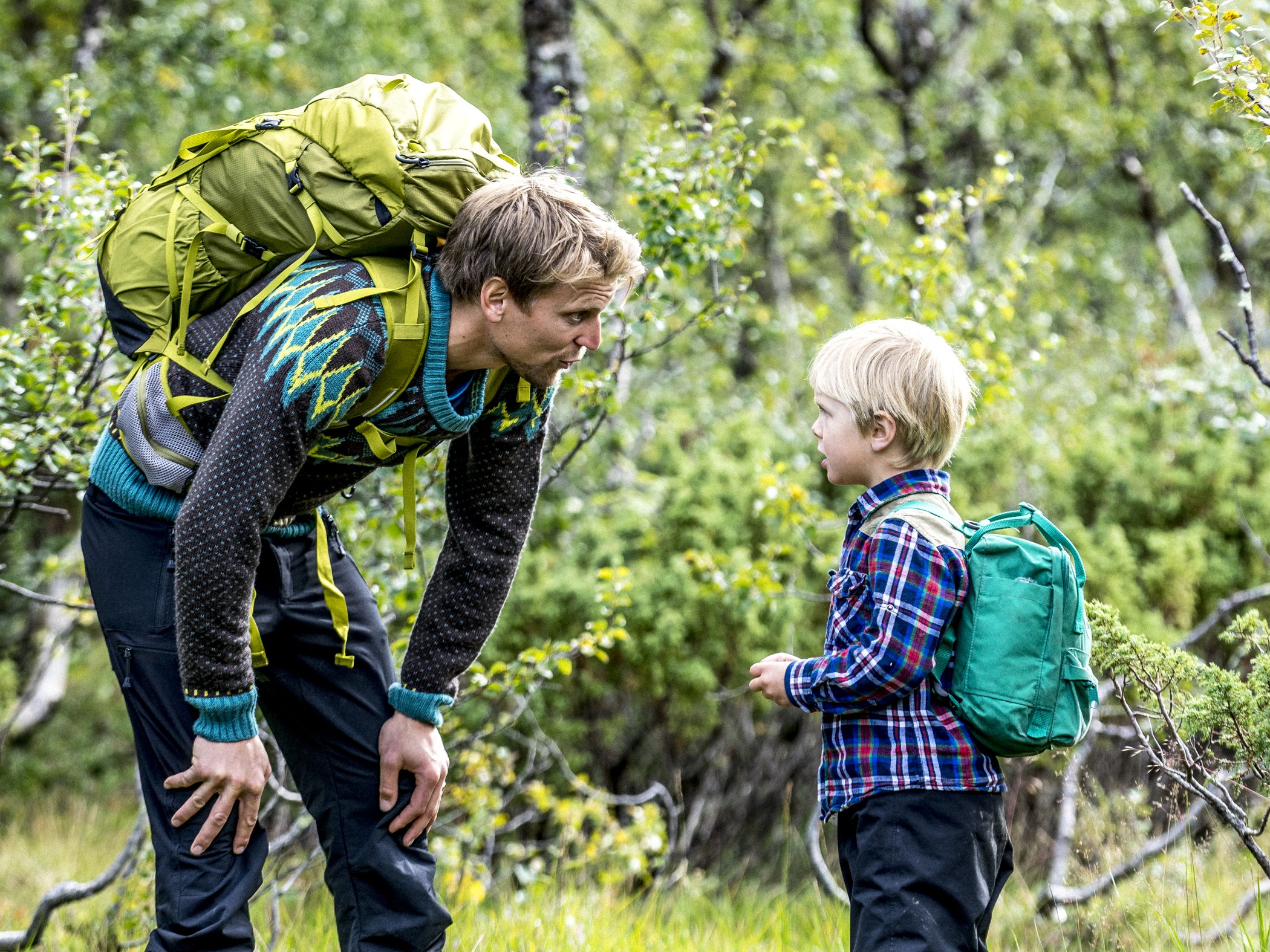 Adult hiker talking with a child carrying a backpack on a forest trail.