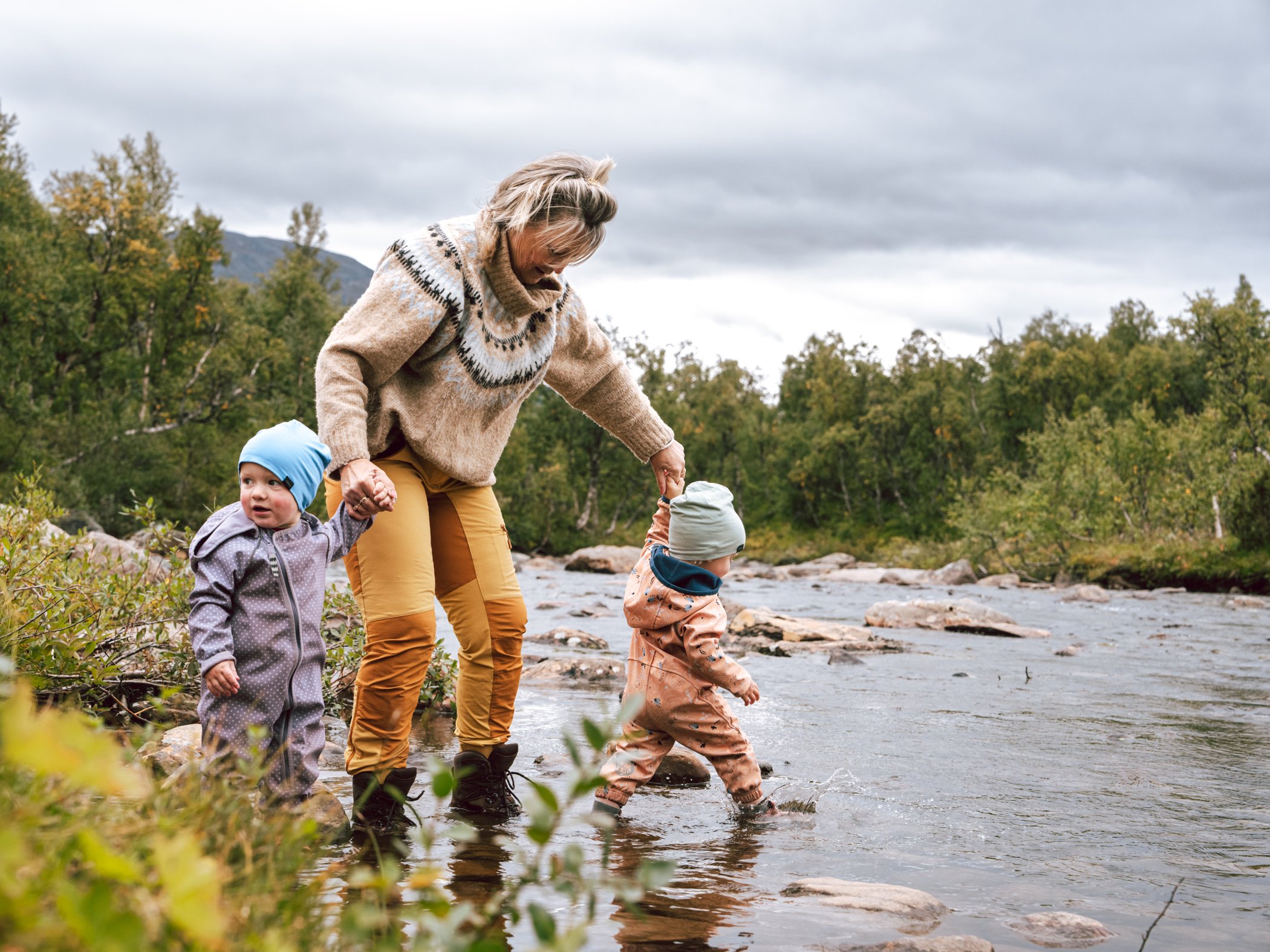 Adult helping two children cross a shallow stream in a mountain landscape.