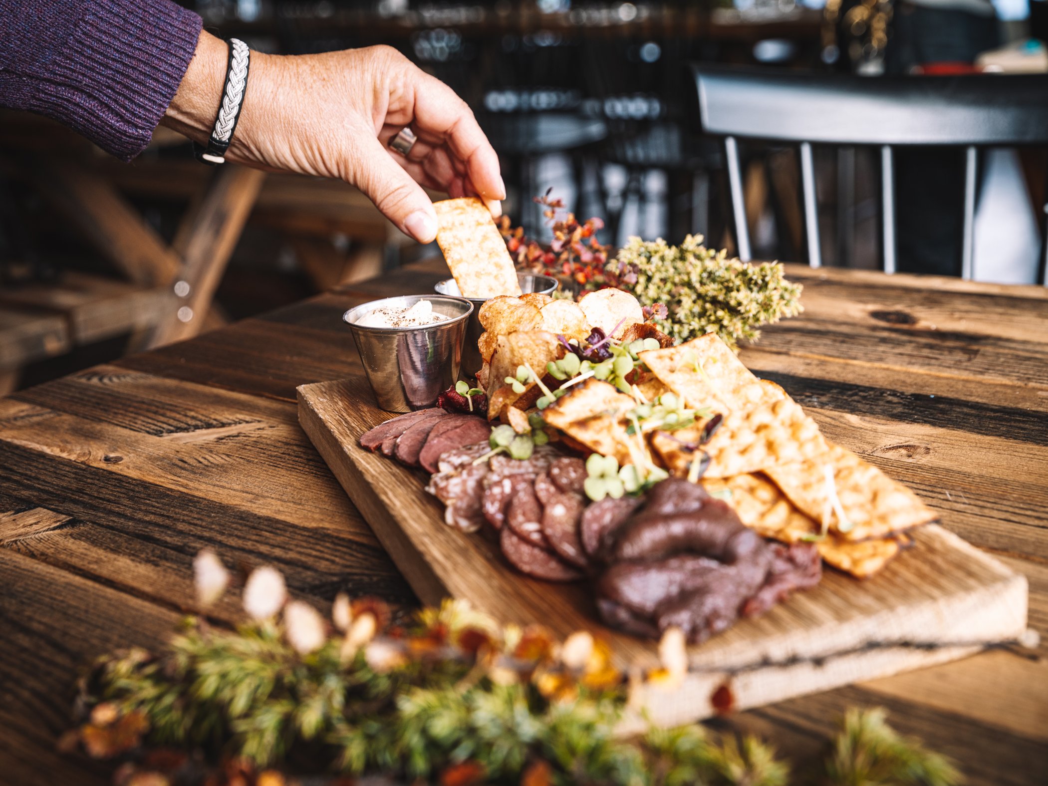 Hand reaching for a piece of flatbread from a mountain-inspired charcuterie board with local delicacies on a wooden table.