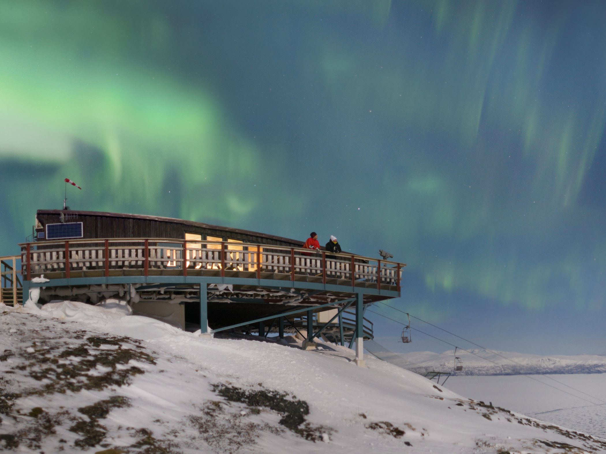 Two people on the terrace at STF Abisko Mountain station as the northern lights illuminate the snow-covered mountain landscape.