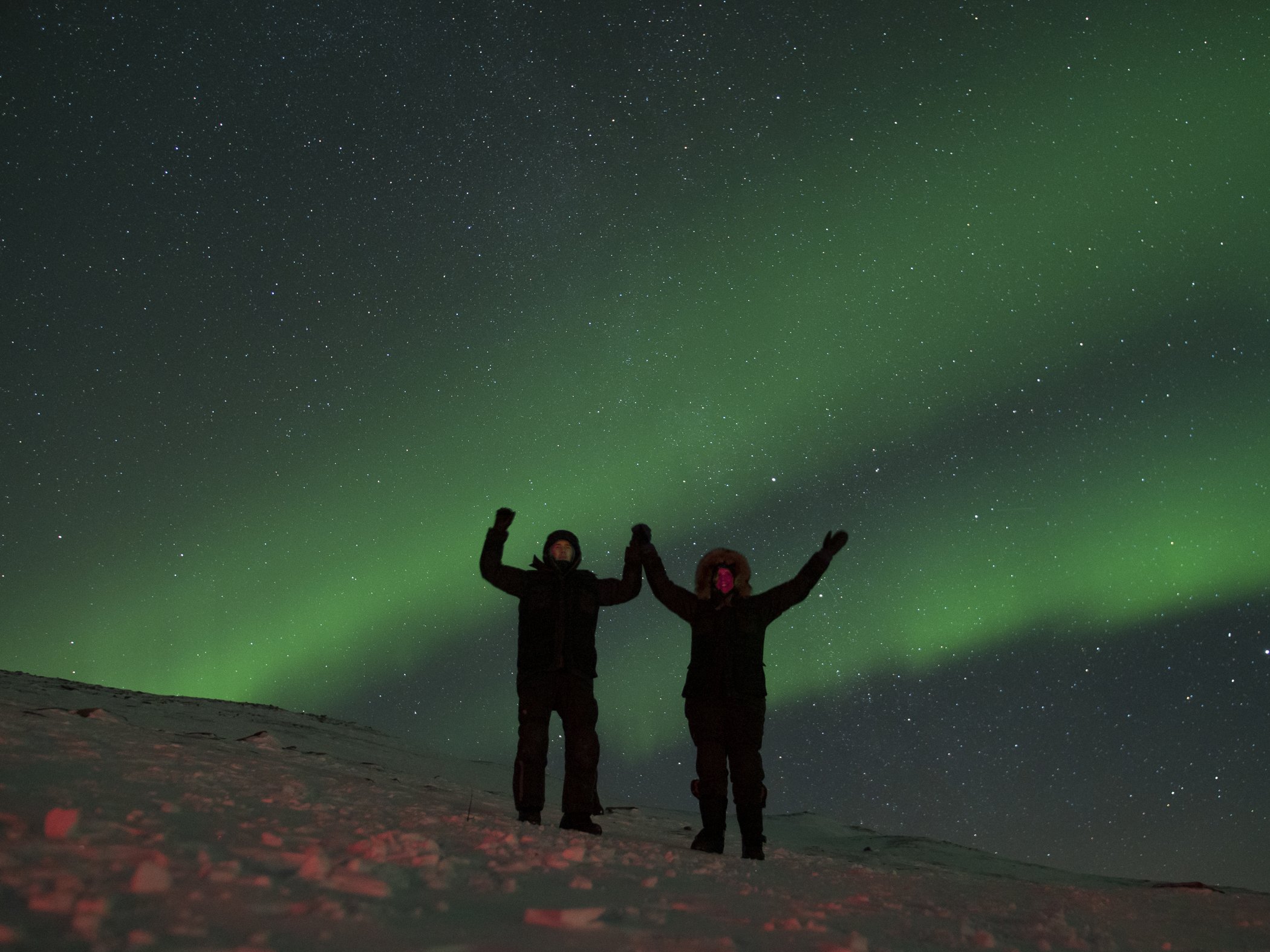 Two people standing in the snow watching the northern lights dance across the starry sky in Abisko.