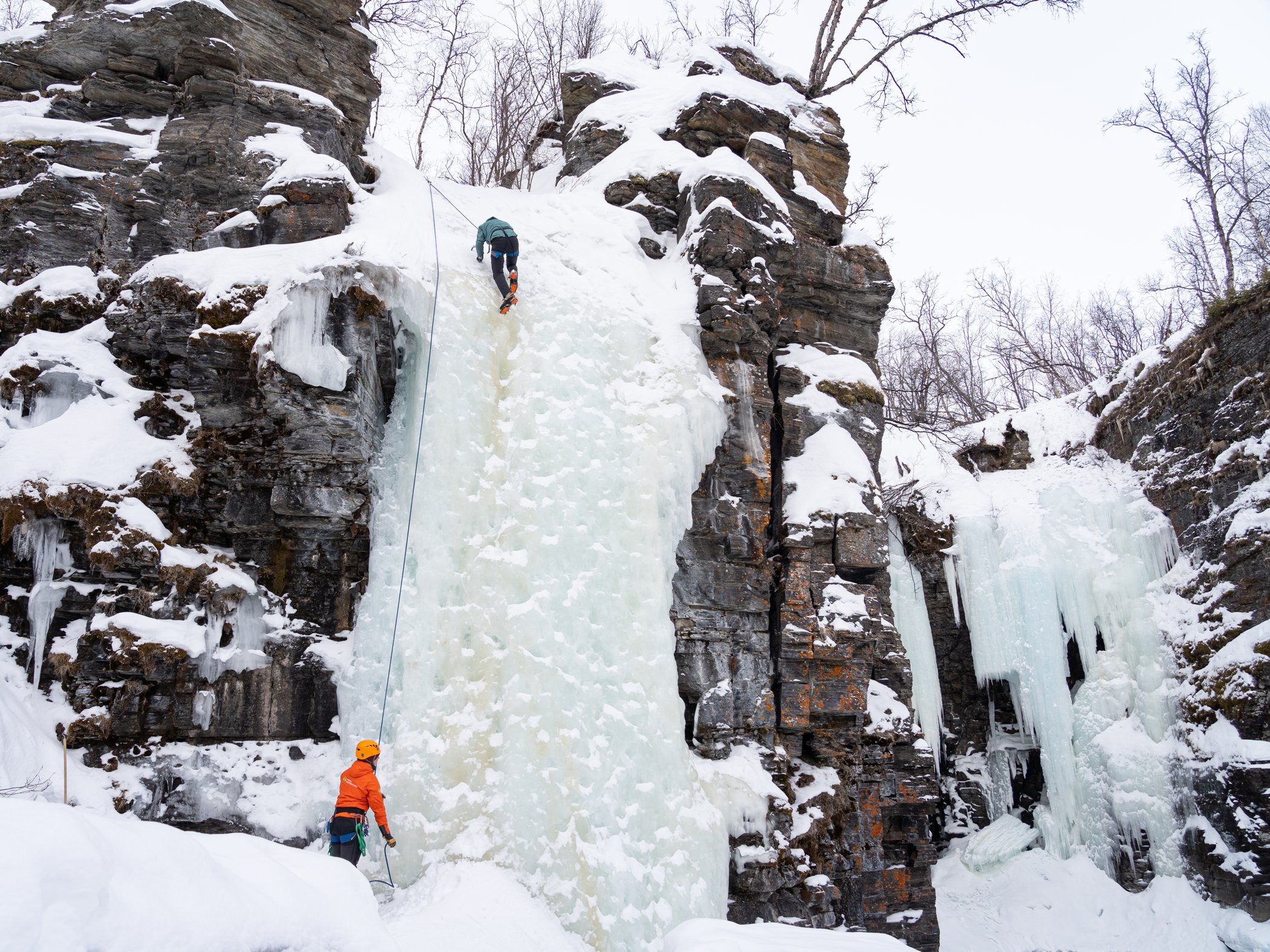 Participant climbs a frozen waterfall while a guide belays from the ground in Abisko.